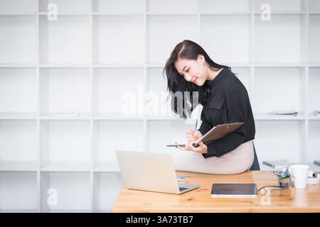 Asian businesswoman sitting on the edge of desk and working in modern office. Stock Photo