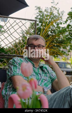 A man relaxes on a patio chair, surrounded by lush green plants and vibrant pink tulips. Stock Photo