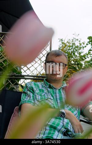 A man relaxes on a patio chair, surrounded by lush green plants and vibrant pink tulips. Stock Photo