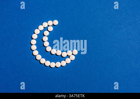 White melatonin pills in shape of crescent moon on dark blue background. Insomnia, healthy sleeping, daily supplements concept. Stock Photo