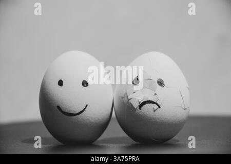 Two chicken eggs, one with joy, and the second broken with sad emotions on the table, sadness and sorrow Stock Photo