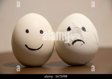 Two chicken eggs, one with joy, and the second broken with sad emotions on the table, sadness and sorrow Stock Photo
