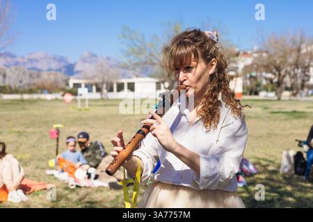 A woman plays a wooden flute in a grassy area at an outdoor festival. She wears a white shirt and has curly hair. In the background, other people enjo Stock Photo