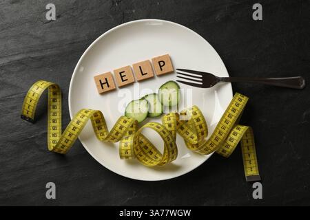 Bulimia. Plate with word Help, pieces of cucumber, measuring tape and fork on dark textured background, top view Stock Photo