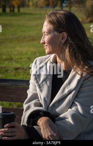 Woman sitting on a park bench, enjoying coffee on a sunny autumn day, surrounded by nature's tranquility. Vertical photo Stock Photo