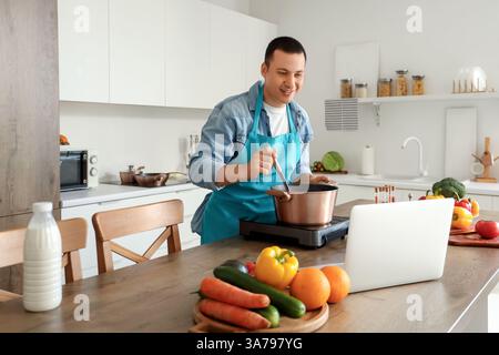 Young man with laptop cooking soup in kitchen Stock Photo