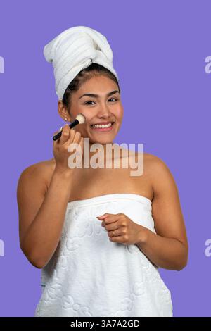 Happy young woman wearing bath towel applying blush with makeup brush after shower, isolated on purple background Stock Photo