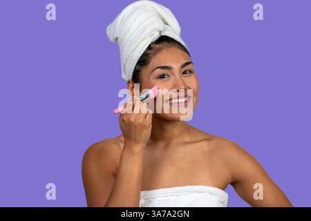 Happy woman wearing towel applying clay mask with brush, enjoying skincare routine Stock Photo