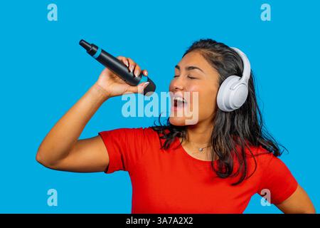 Young latin woman enjoying singing with wireless microphone and headphones, isolated on a blue background Stock Photo