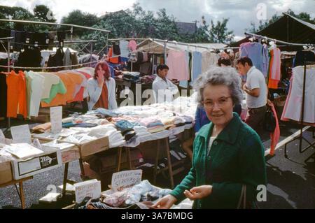 A lively snapshot from a bustling 1960s British street market, captured on 35mm colour slide film. The image features a smiling woman browsing a clothing stall filled with brightly coloured garments and handwritten signs, surrounded by classic market scenes and local shoppers. Stock Photo