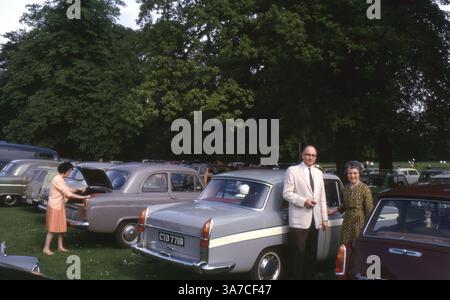 A man and woman pose for a photo beside a grey Austin Cambridge in a parkland car park in England, June 1965. The scene captures a typical mid-1960s summer outing, with families unloading cars and preparing for the day. Stock Photo