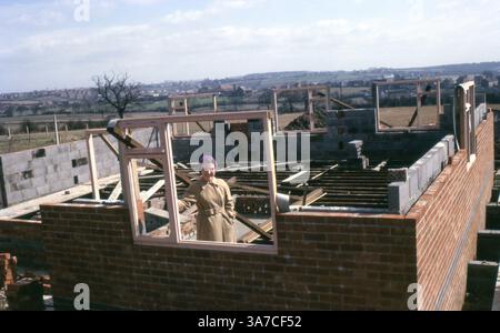 A woman stands proudly at the construction site of her future home, captured in June 1965. Surrounded by countryside and brickwork, the image shows the early phases of housebuilding in 1960s Britain, including timber window frames and concrete floor joists. Stock Photo