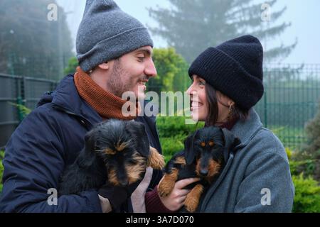 Young couple holding their two wire-haired dachshunds smiling at each other in a cold winter day Stock Photo
