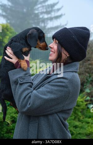 Young woman with winter clothes holding her dachshund dog, smiling and looking at him in a foggy day Stock Photo