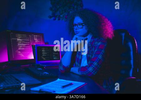 Young female programmer working remotely on her computer in cyberspace, analyzing code within a vibrant and professional home workplace. Stock Photo