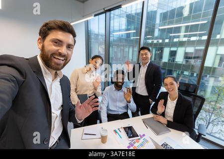 A diverse group of colleagues in a modern office wave to the camera, radiating positivity during a video call or team selfie. Stock Photo
