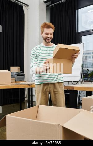 A man happily unpacks a box in a modern office filled with empty boxes and supplies. Stock Photo