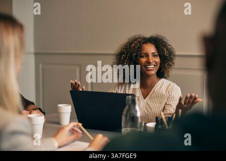 Happy curly haired young businesswoman sitting with laptop while discussing during meeting in office Stock Photo