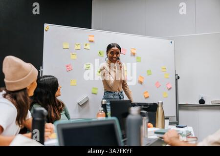Happy young female student standing near whiteboard while discussing with friends during project meeting in classroom at Stock Photo