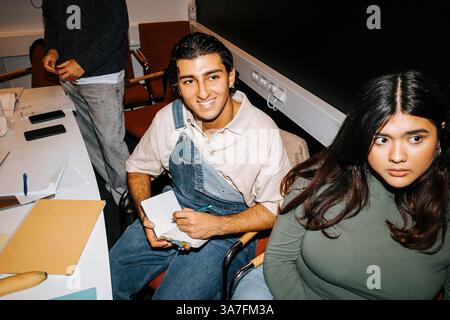 High angle view of smiling young man wearing bib overalls taking notes while sitting near female friend in classroom at Stock Photo