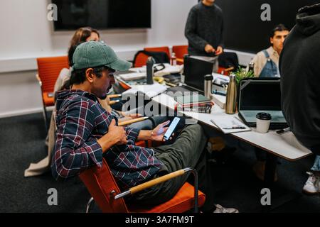 Man wearing cap using smart phone while sitting in classroom with friends at college Stock Photo