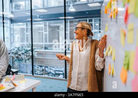 Happy mature female teacher standing near whiteboard while teaching in tech university Stock Photo