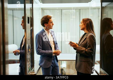 Male and female business professionals talking with each other while standing in elevator at office Stock Photo