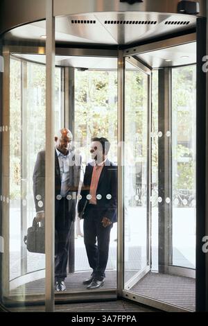 Happy male business colleagues walking through revolving door of office building Stock Photo