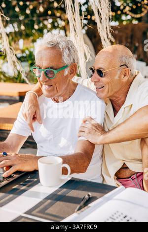 Happy senior gay man sitting with arm around boyfriend using laptop Stock Photo