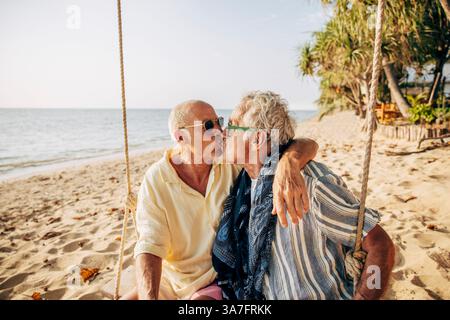 Affectionate senior gay couple kissing while sitting on rope swing at beach Stock Photo