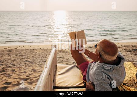 Relaxed senior man lying on bed while reading book on beach at sunset Stock Photo