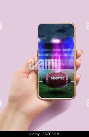 Person holding smartphone with American football ball on stadium and filled stands in the background during open air match event. Composite image Stock Photo