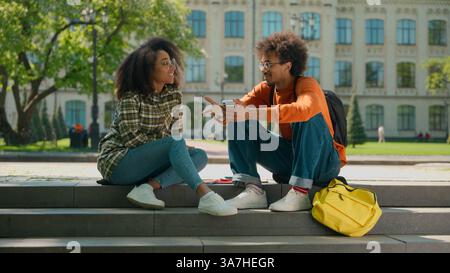 Two happy African American friendly students conversation sitting outdoor multiracial friends African American woman girl female with boy man guy in Stock Photo