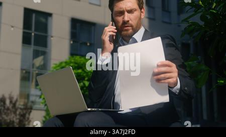 Multitasking busy Caucasian middle-aged businessman business man male guy entrepreneur in city talking mobile phone call using laptop working job Stock Photo