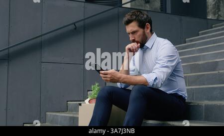 Serious fired unemployed Caucasian middle-aged businessman using smartphone searching work vacancy online thinking business man male guy in city with Stock Photo