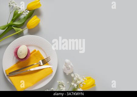 Spring Easter table setting with yellow tulips, white plate, bunny, eggs on gray background. Happy Easter celebration. Copy space. View from above. Stock Photo
