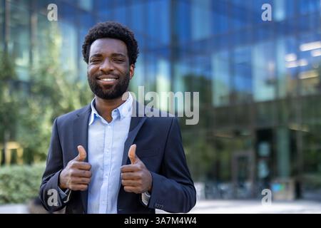 A young, cheerful businessman expresses positivity with a thumbs-up gesture in front of a modern building on a sunny day. Stock Photo