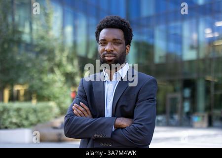 A confident Black businessman stands with crossed arms in front of a modern office building with blurred greenery. Stock Photo