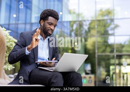 A confident African-American businessman attends a virtual meeting on his laptop outdoors, greeting colleagues and making notes in a notebook. Stock Photo