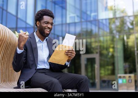 A young businessman cheers with his fist raised while sitting outside, having just opened a letter with great news in front of an office building Stock Photo