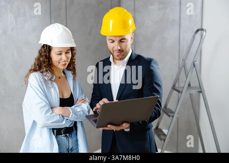 Attractive young woman contractor is talking to customer in helmet, and demonstrates sequence of repairs in living room on laptop. Meeting of landman Stock Photo