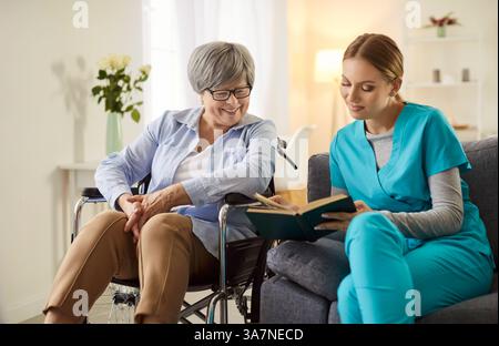 Young caregiver or social worker reading a book for senior woman with disability in wheelchair. Stock Photo