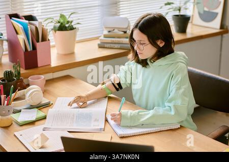 Student with prosthetic hand studying with books, plants, and gadgets on wooden desk while taking notes in cozy workspace in daylight with bright surroundings Stock Photo