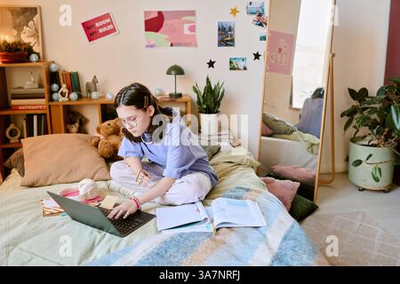 Brunette teenager with prosthetic hand studying on bed in cozy bedroom filled with books, plant, and mirror reflecting some parts of the room Stock Photo
