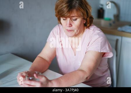 Thoughtful senior woman sitting at kitchen table with a glass mug, reflecting in quiet moment. Stock Photo