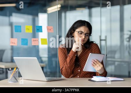 Serious thinking woman using tablet computer. Businesswoman multitasking working inside office with notebook. Concentrated female employee reading electronic report online email. Stock Photo