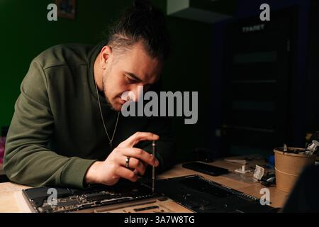 Portrait of technician male carefully using screwdriver to reassemble laptop after performing repairs on motherboard in specialized workshop with dark Stock Photo