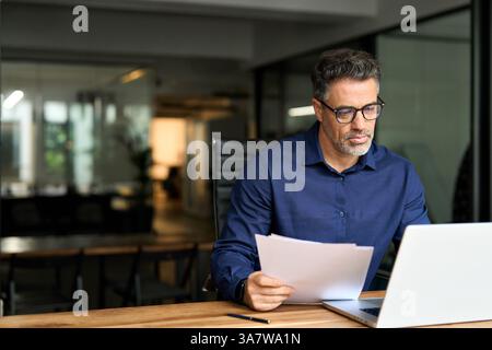 Busy mature professional business man using laptop reading document at work. Stock Photo