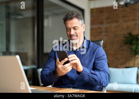 Smiling mature business man corporate leader using cellphone sitting in office. Stock Photo