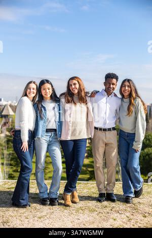 Five multi ethnic students standing together in a park with arms around each other, smiling Stock Photo